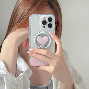 A person holding a smartphone with a clear phone case featuring a pink heart-shaped ring stand, taking a mirror selfie.