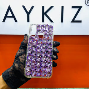 A hand holding a mobile phone case adorned with purple rhinestones, with the brand name AYKIZ visible in the background.