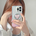 A person holding a smartphone with a clear phone case featuring a pink heart-shaped ring stand, taking a mirror selfie.